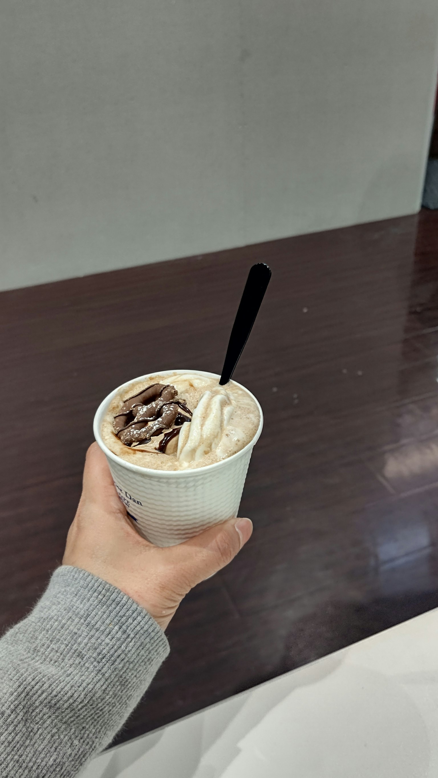 Tangan memegang cangkir es krim dengan sendok hitam di atas meja kayu