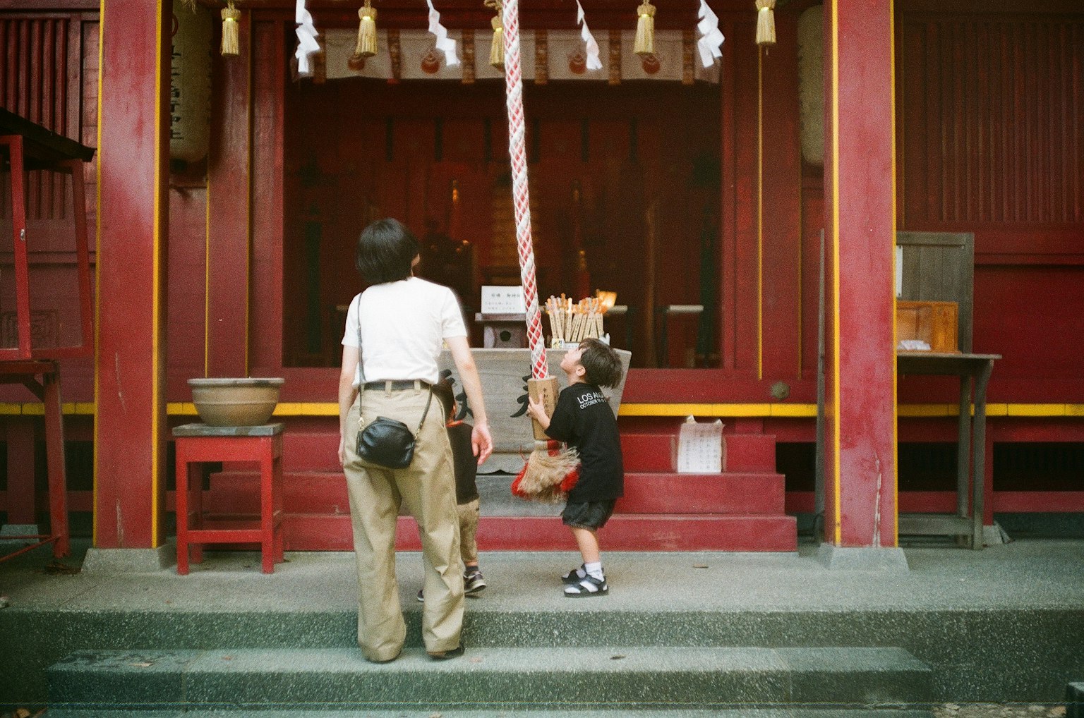 เด็กเล่นกับเชือกหน้าศาลเจ้ากับผู้ใหญ่ ผนังสีแดงและการตกแต่งที่โดดเด่น