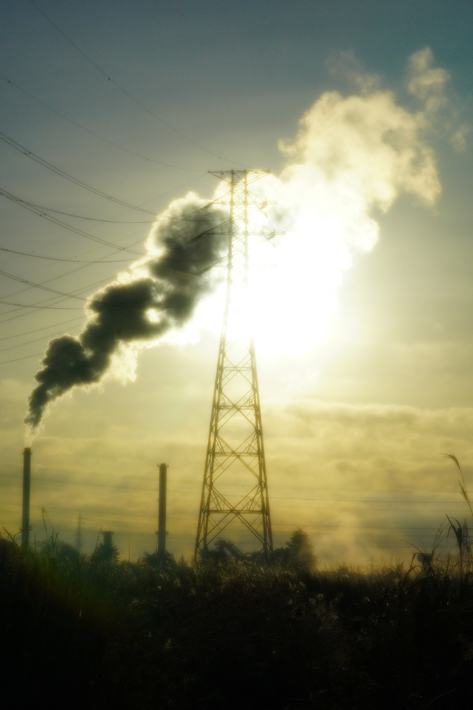 Silhouette di una torre elettrica e camini che emettono fumi contro un tramonto