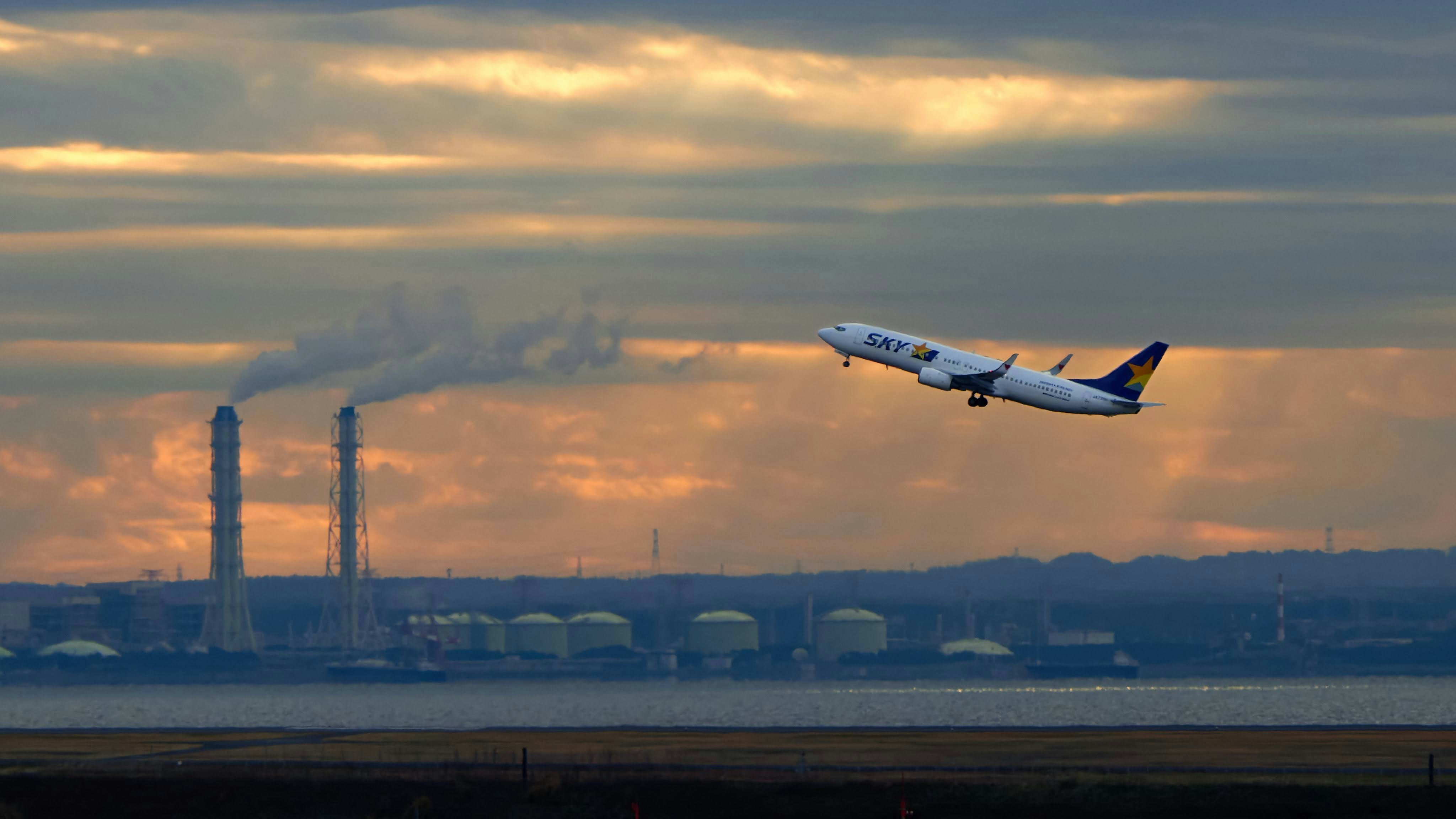 工場の煙突と水辺を背景に飛行機が飛ぶ夕景