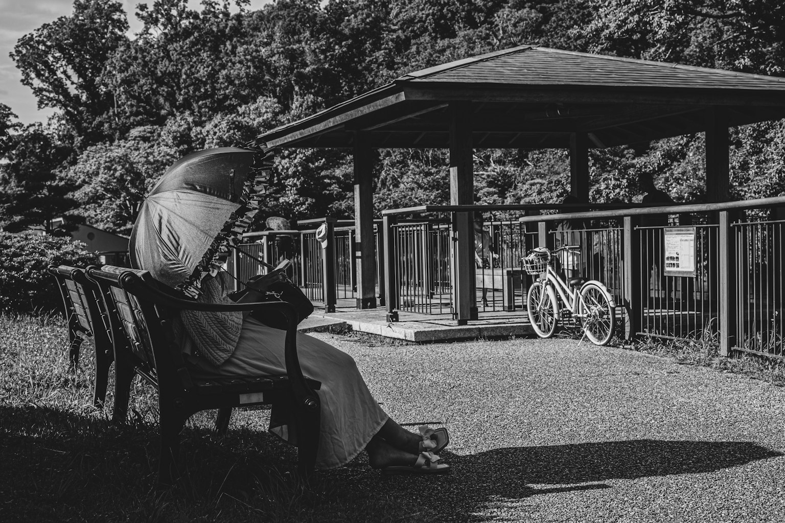 一位女性坐在公園長椅上撐著陽傘 背景有自行車和涼亭