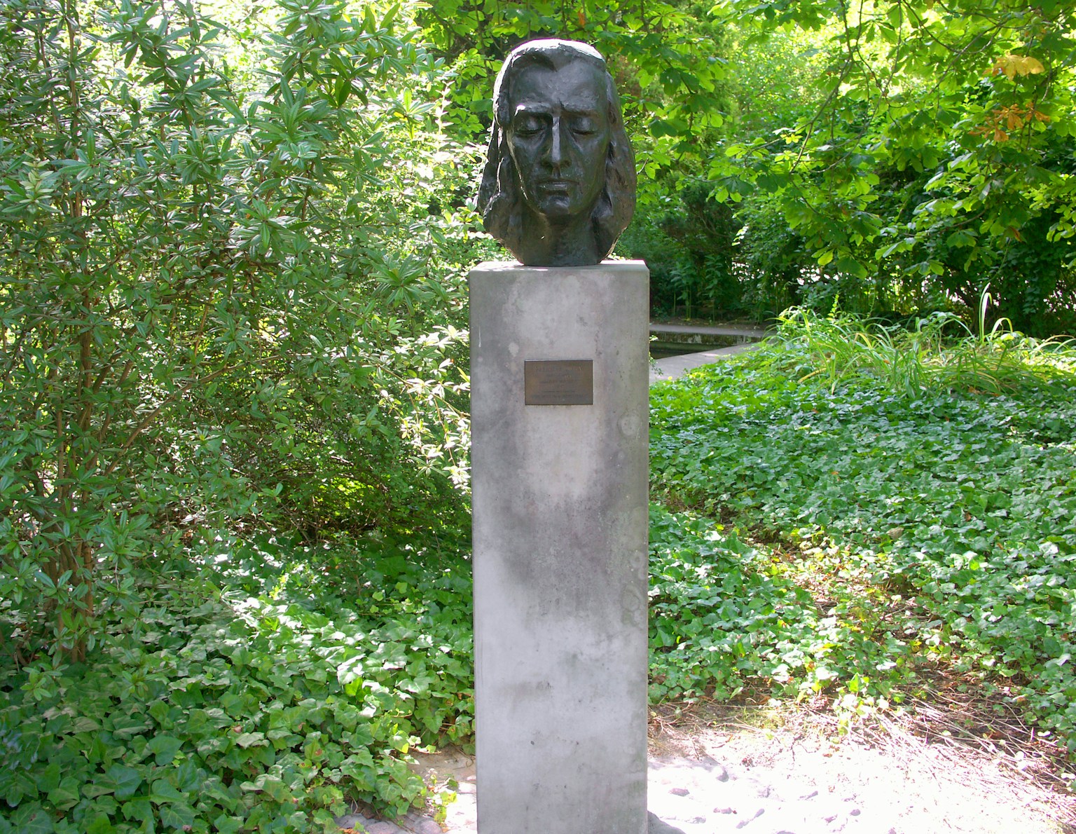 Monumen dengan patung kepala perunggu dikelilingi tanaman hijau