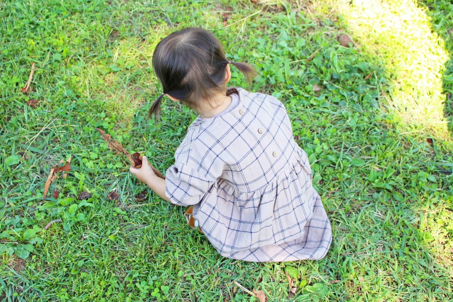 Anak duduk di atas rumput bermain dengan tongkat mengenakan gaun bermotif