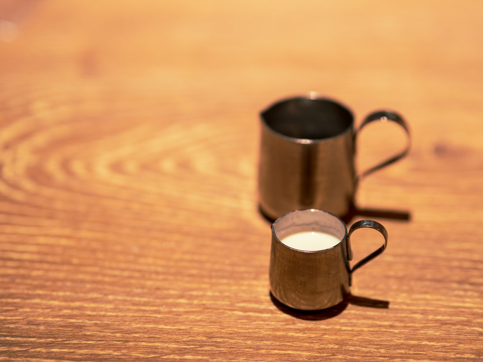 Un petit pichet à lait en métal à côté d'un plus grand pichet à lait en métal sur une table en bois