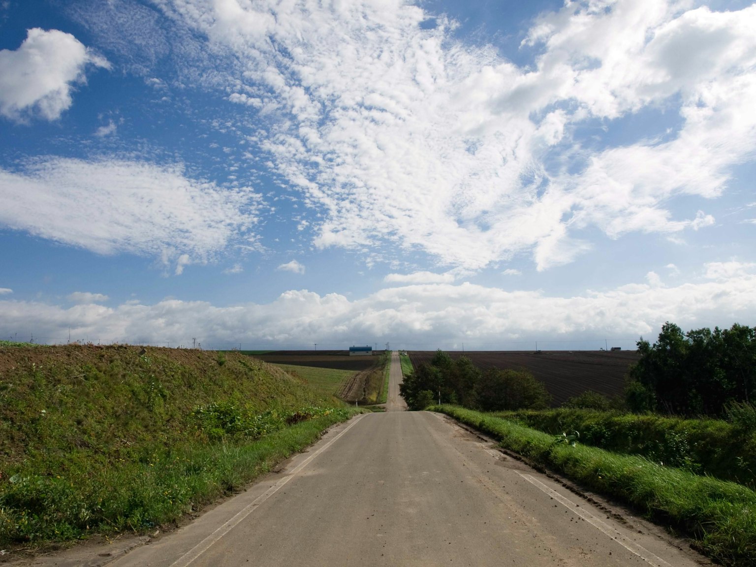 Jalan beraspal yang membentang ke lanskap dengan langit biru dan awan putih