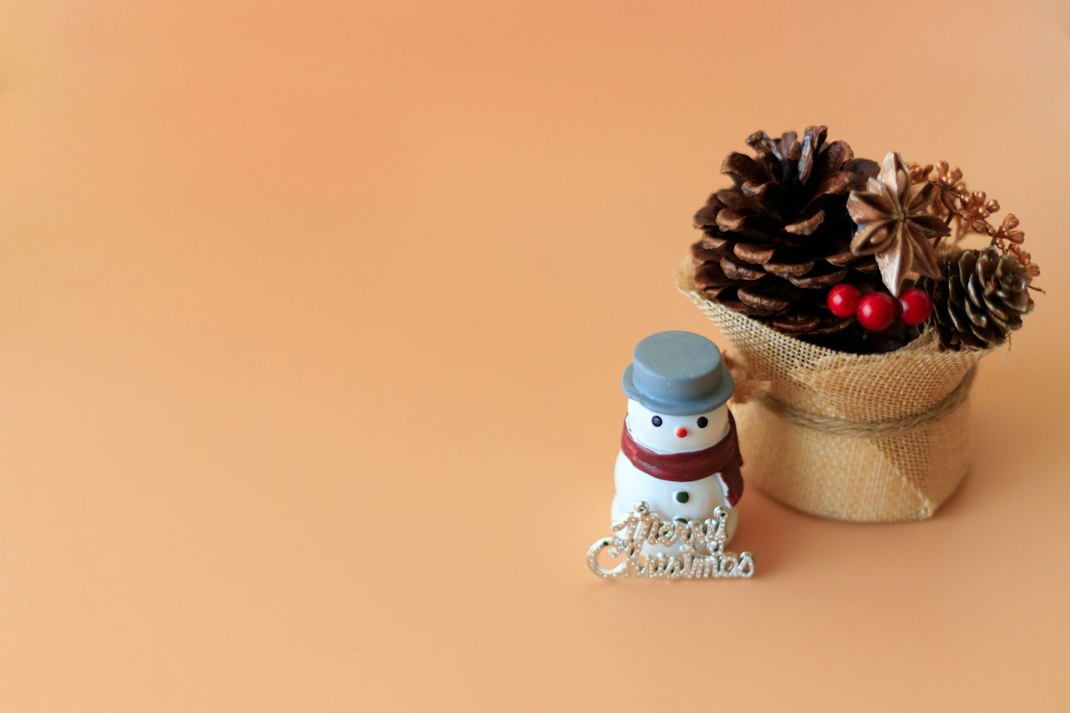 Una figura de muñeco de nieve junto a una maceta con piñas y bayas rojas sobre un fondo naranja