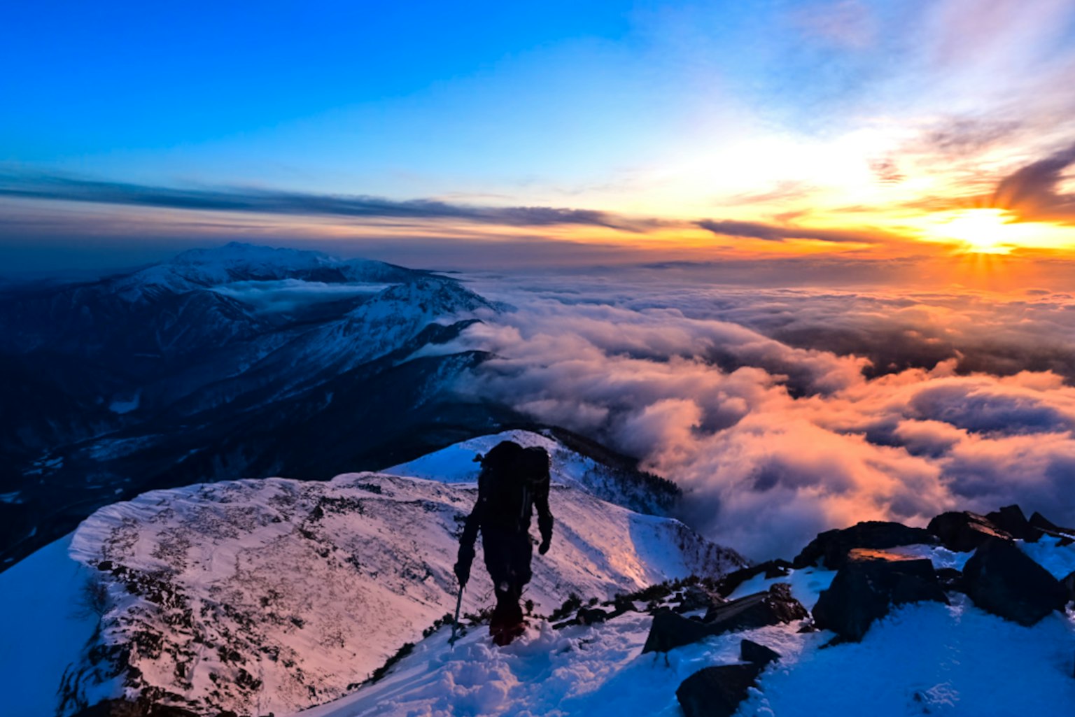 Un grimpeur marchant sur un sommet enneigé avec un coucher de soleil vibrant et des nuages en dessous