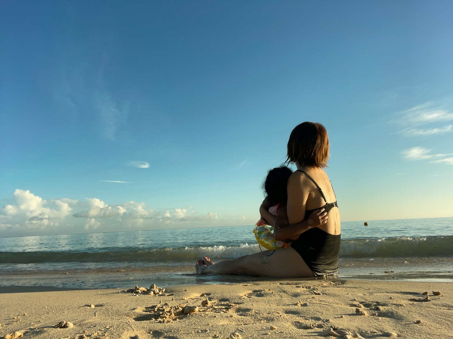 Silhouette einer Frau, die ein Kind am Strand hält, mit blauem Himmel und Ozean im Hintergrund