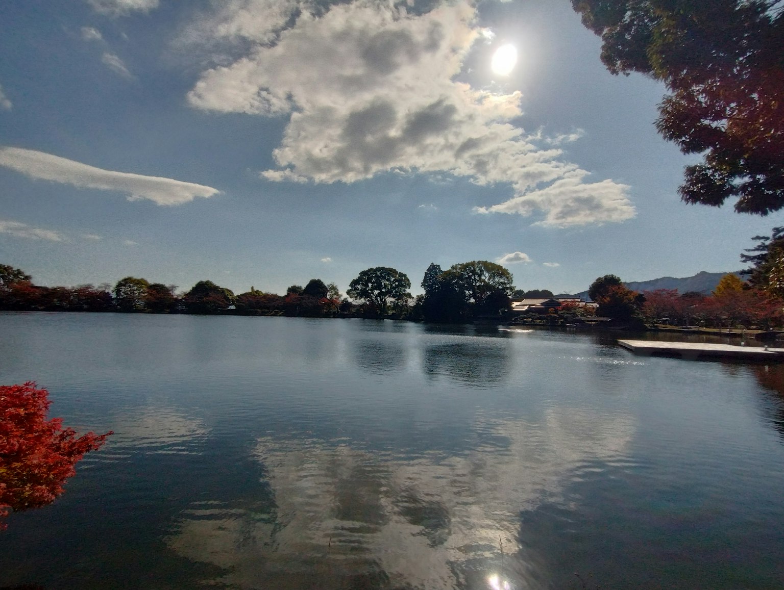 Ruhiger See, der den blauen Himmel und die weißen Wolken spiegelt Rote Blätterbäume am Ufer