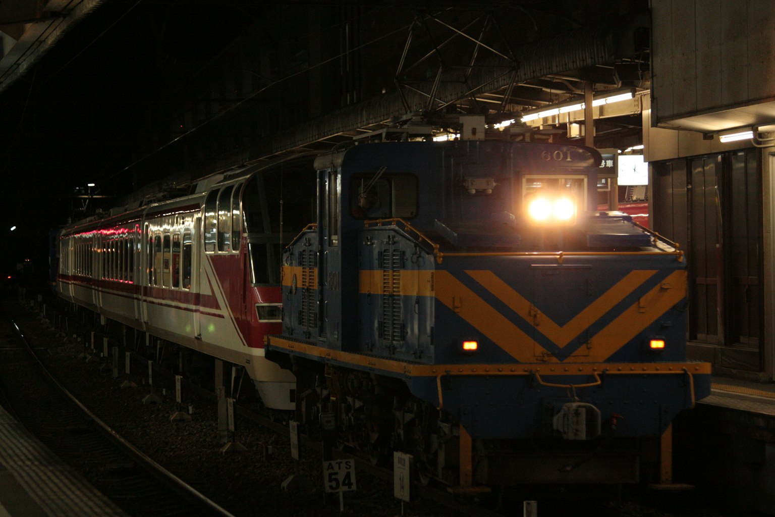 A blue and orange locomotive at a station with a red train during nighttime