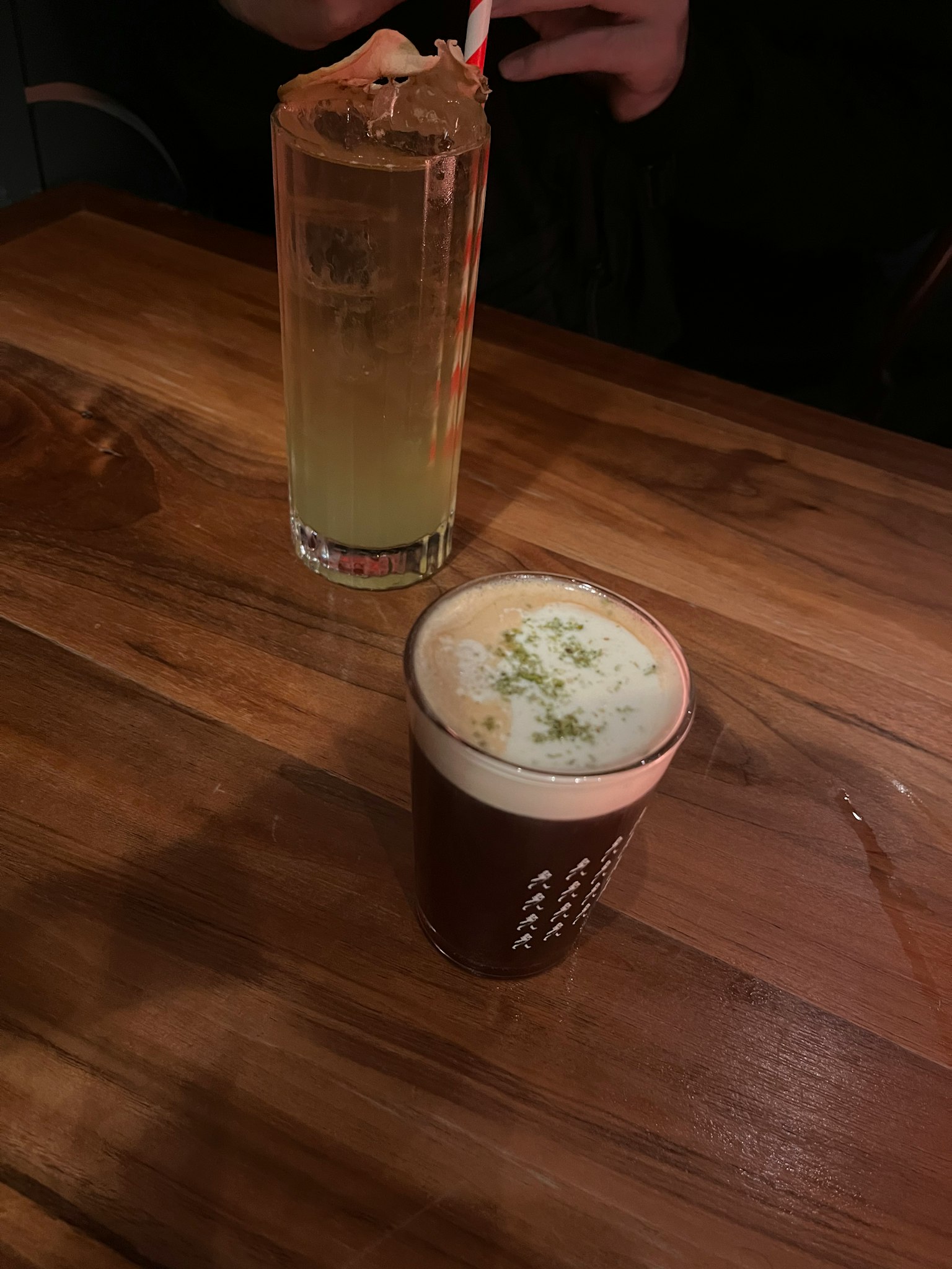 Two drinks on a wooden table one in a tall clear glass another in a brown cup with foam