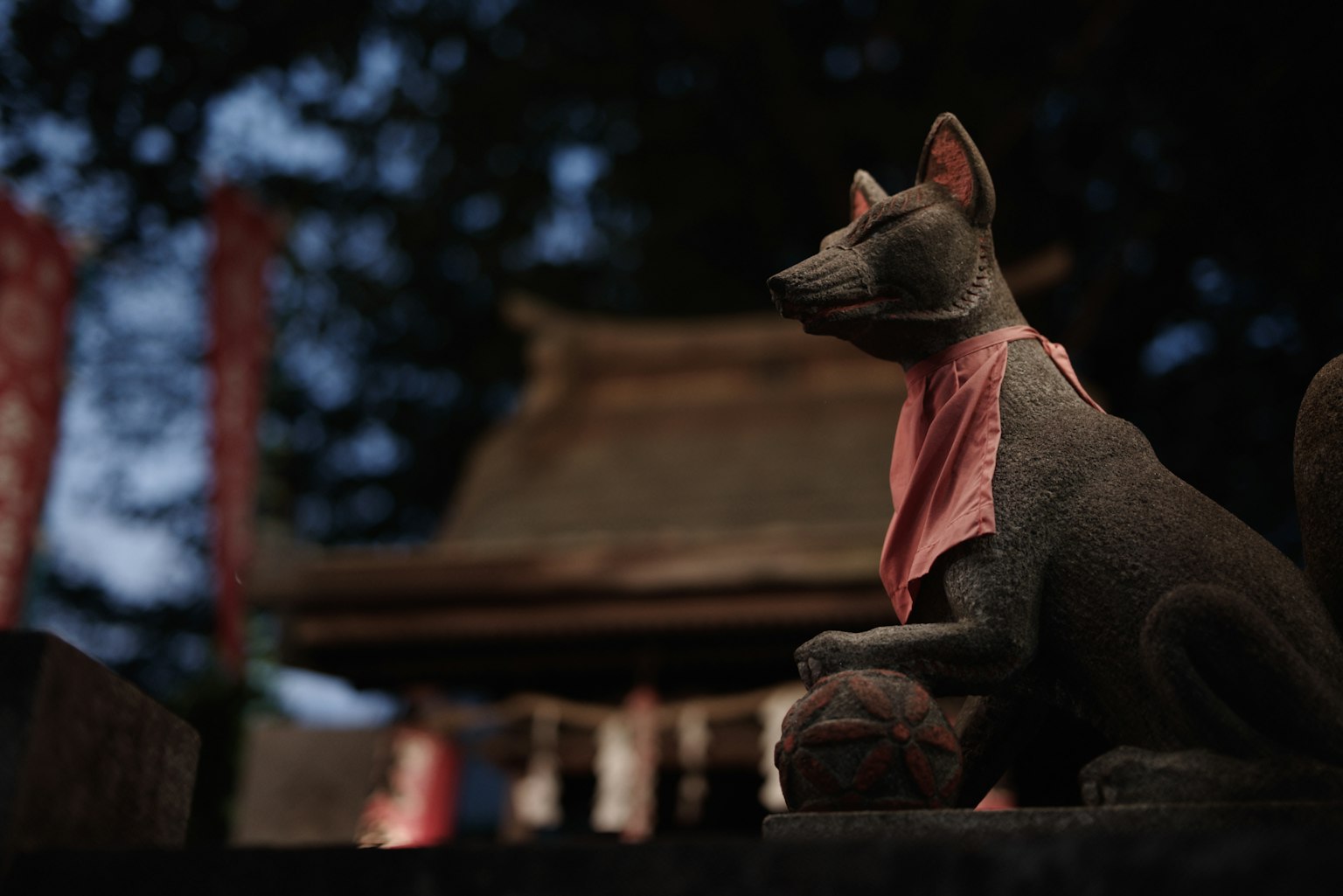 Una estatua de zorro en un santuario que lleva una bufanda roja con un fondo borroso