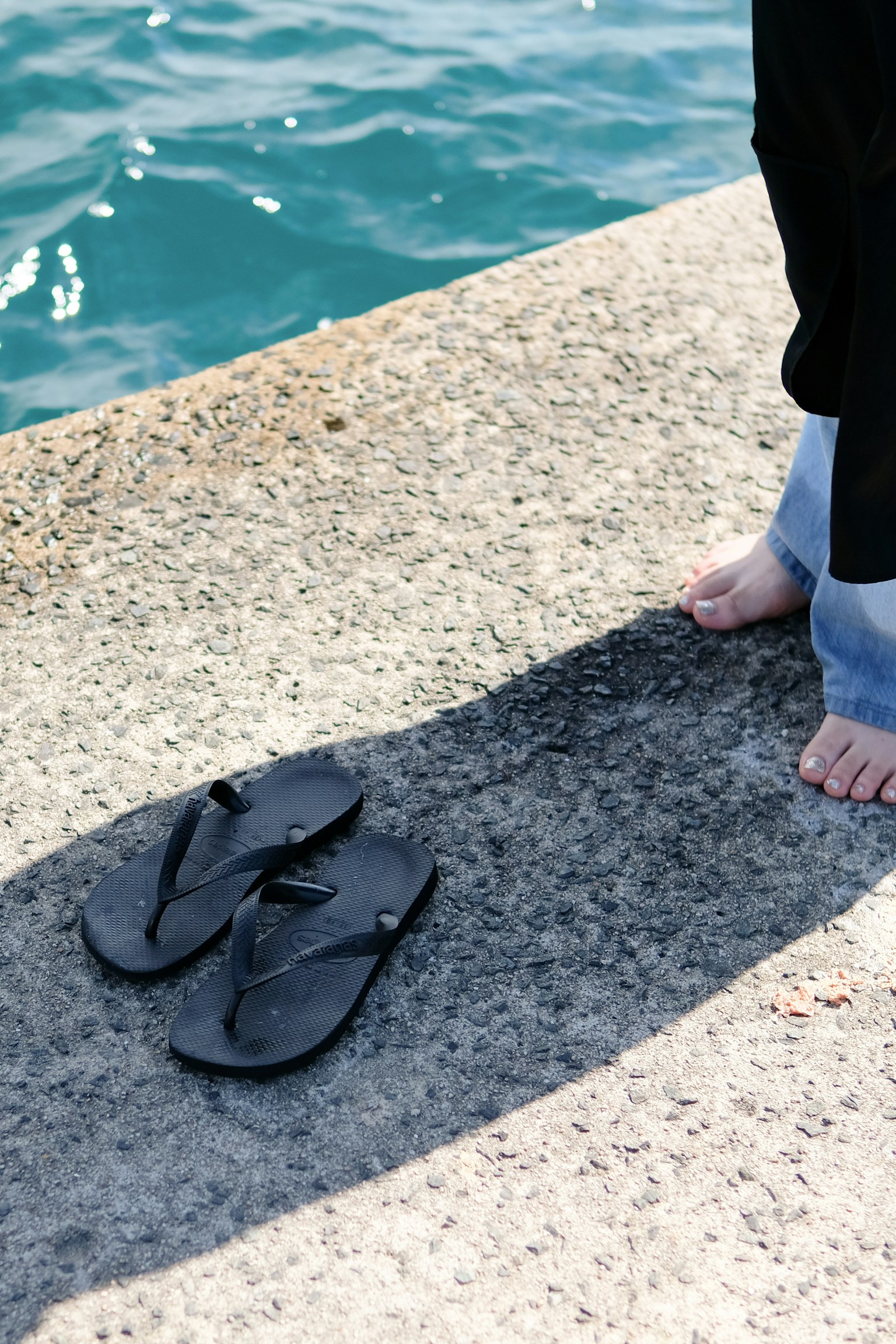 Barefoot person standing next to black flip-flops by the water