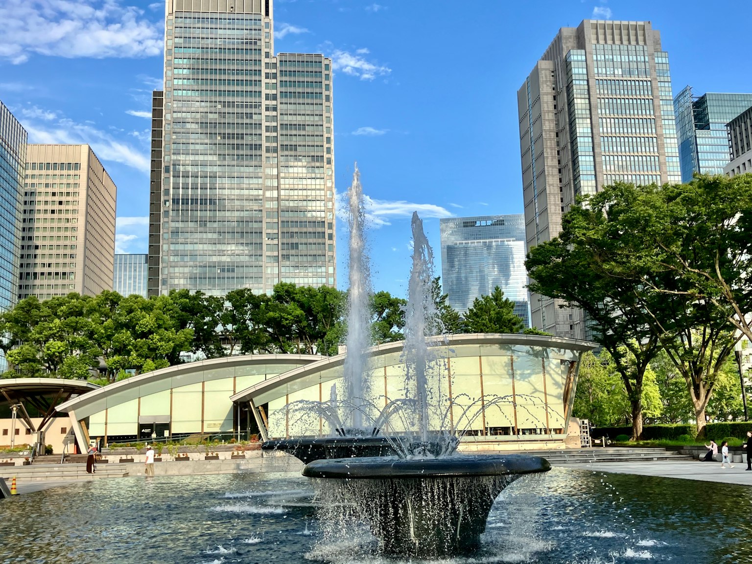 Pemandangan air mancur dikelilingi oleh gedung pencakar langit dan pohon hijau di sebuah taman