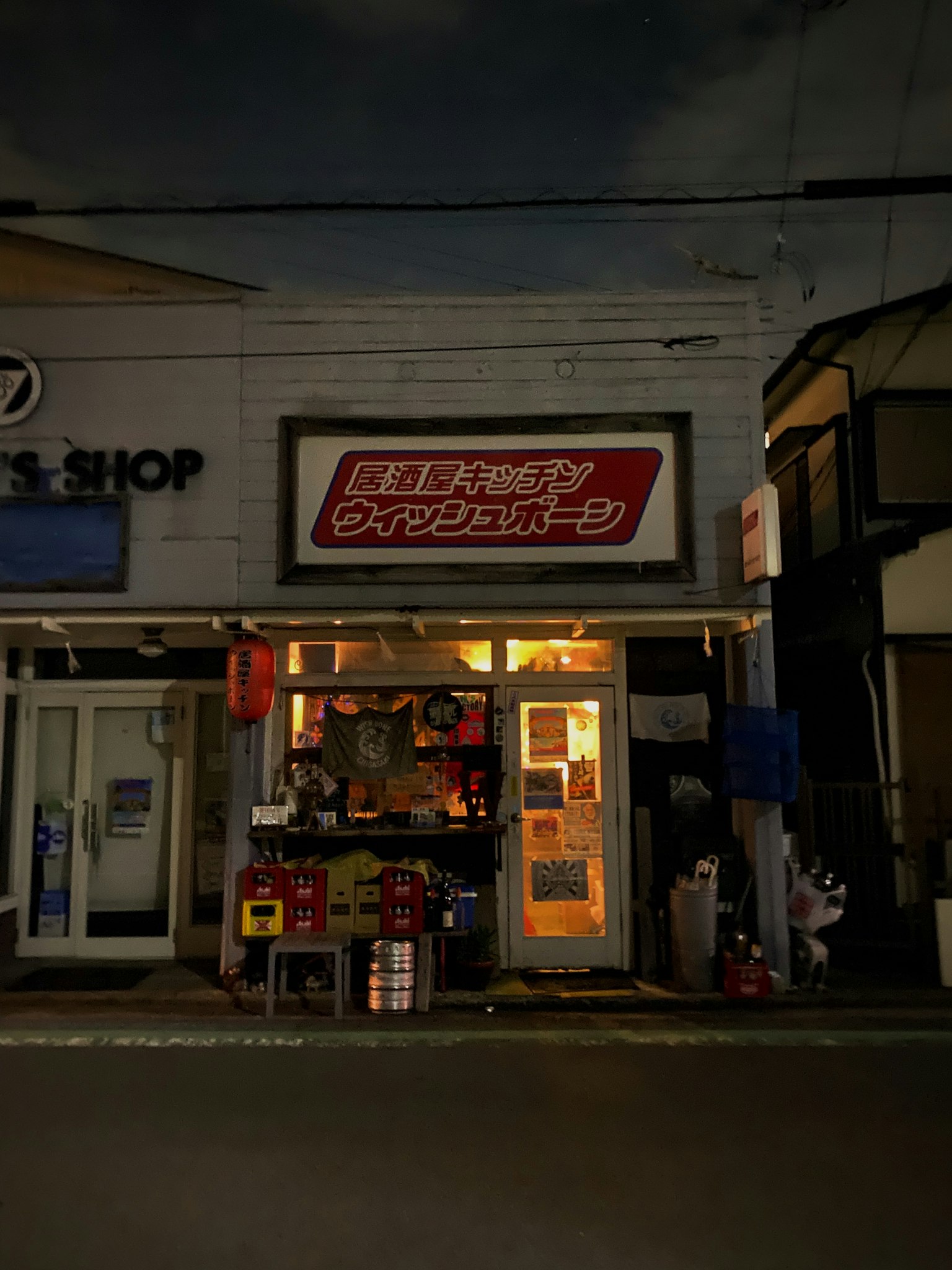 ภาพภายนอกของร้านเล็กๆ ในฉากถนนยามค่ำคืนพร้อมป้ายภาษาญี่ปุ่น