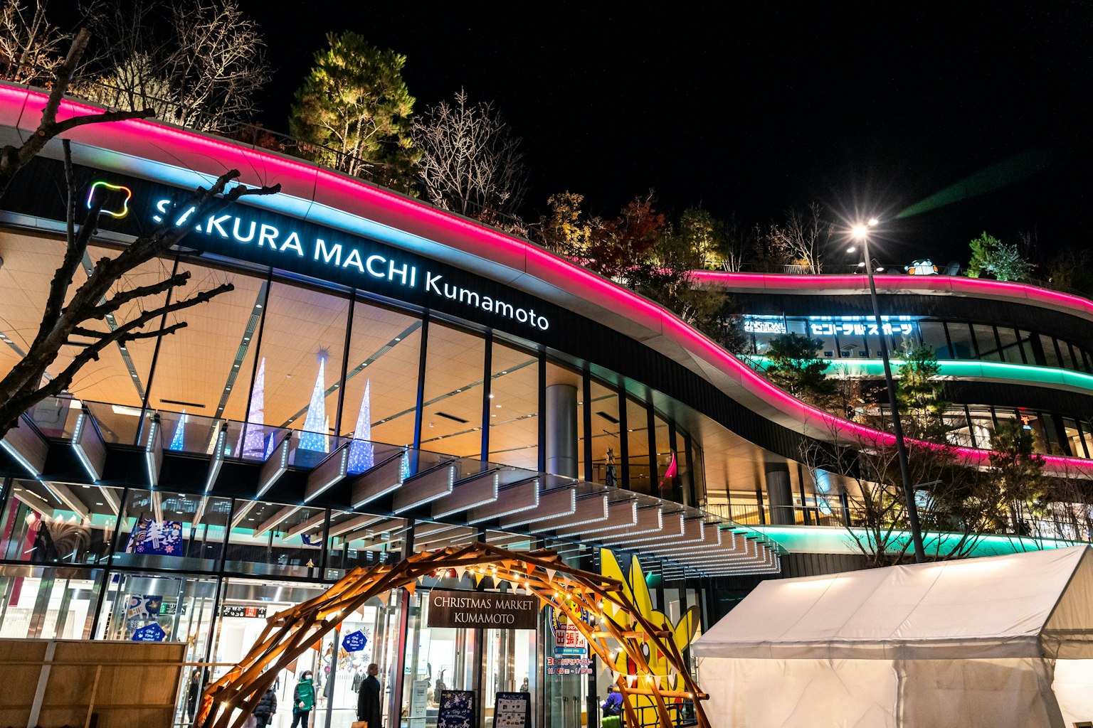 Bâtiment moderne de Sakura Machi Kumamoto la nuit avec des lumières néon brillantes et des arbres environnants