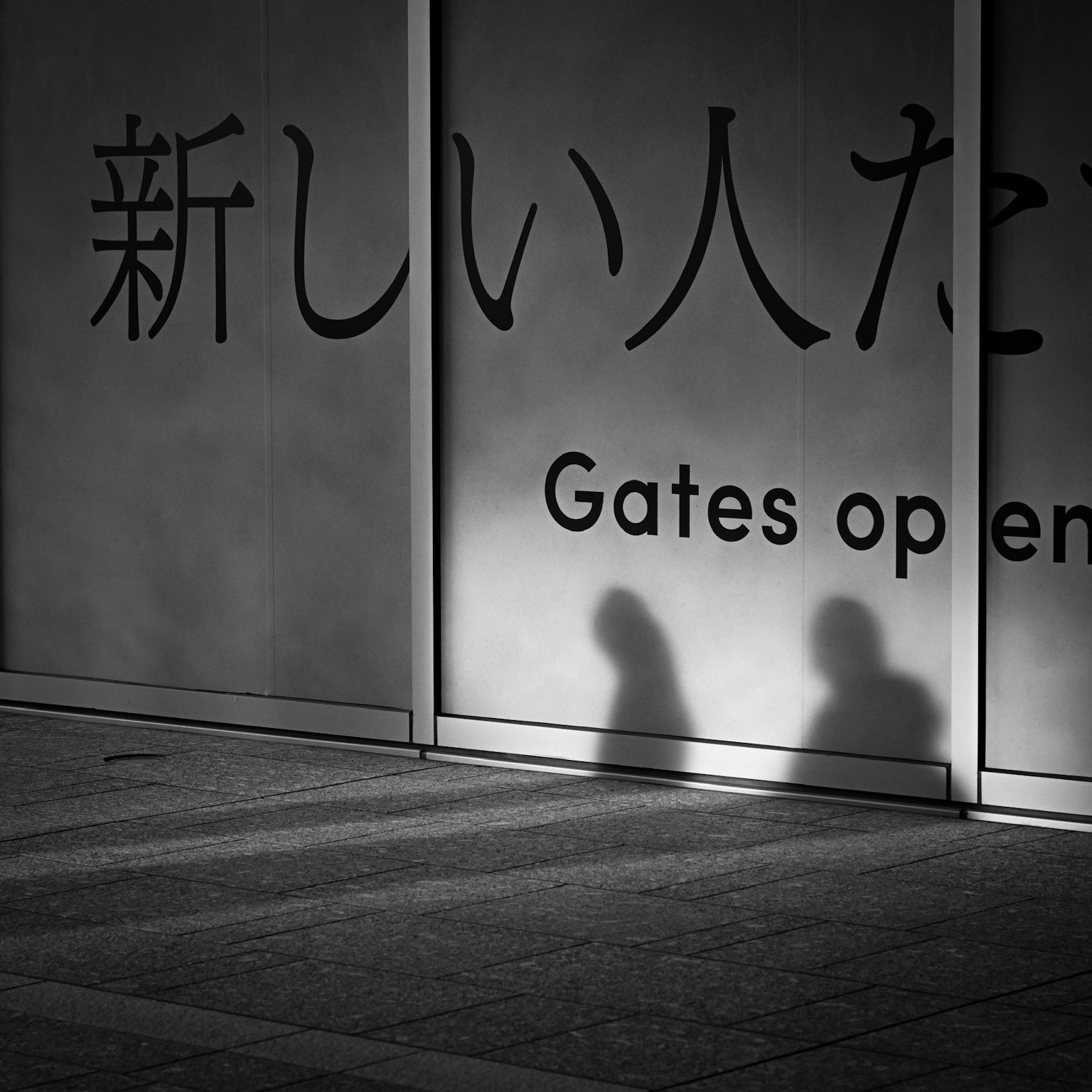 Bayangan dua orang di dinding dengan teks Jepang dan frasa Gates open