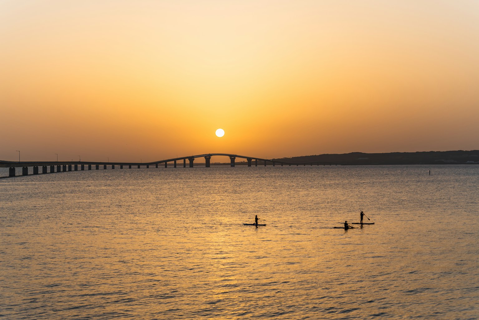 Silhouetted paddleboarders on the ocean at sunset with a bridge in the background