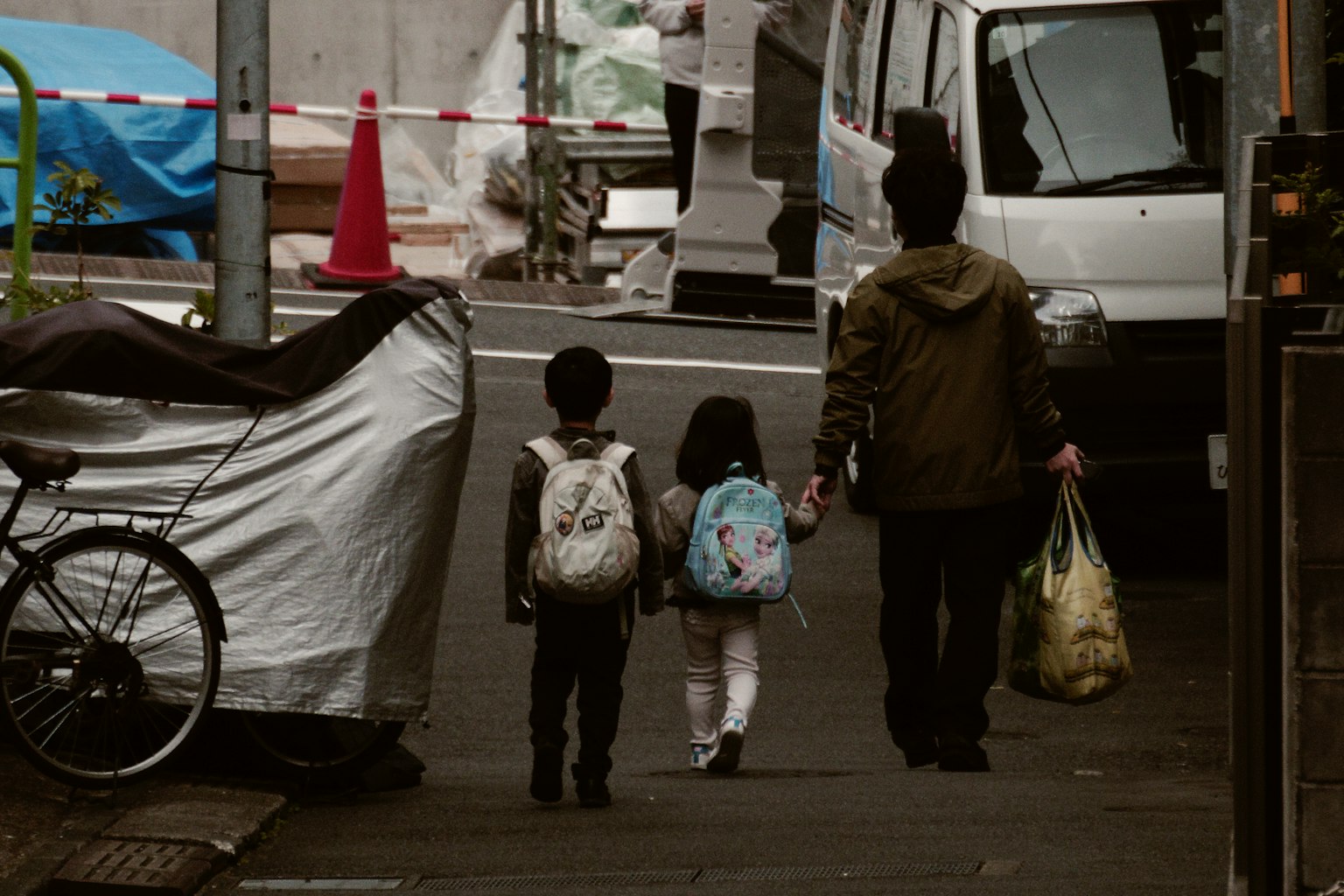 Due bambini e un adulto che camminano mano nella mano in un contesto urbano con un camion e lavori in corso sullo sfondo