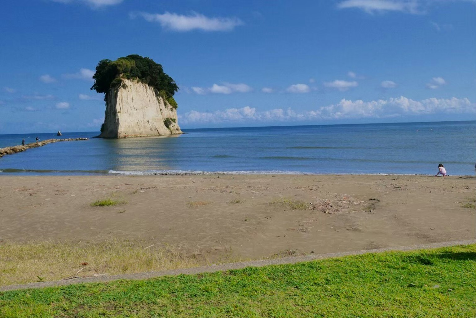 Malersiche Aussicht auf einen Sandstrand mit grünem Gras unter einem klaren blauen Himmel mit einem großen Felsen mit Bäumen
