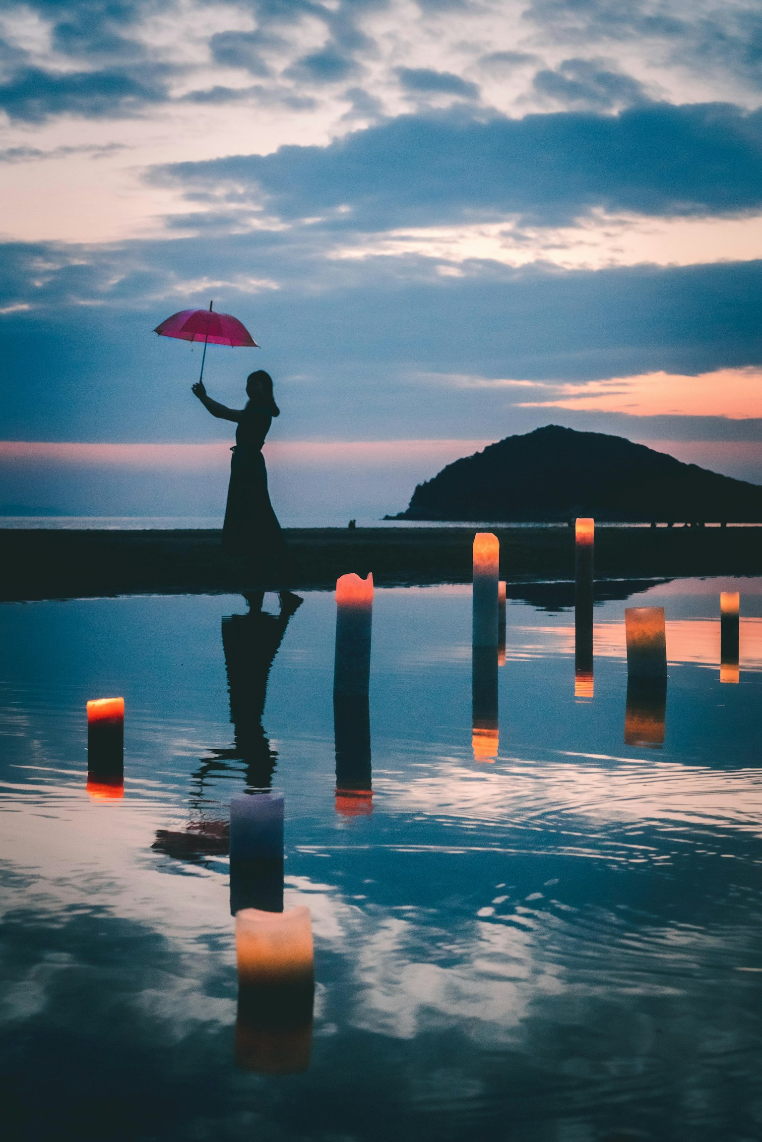 Eine Frau mit einem rosa Regenschirm, die am Wasser entlang geht, umgeben von leuchtenden Kerzen bei Sonnenuntergang