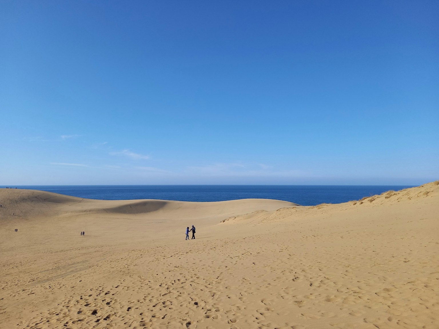 Extensas dunas de arena con un cielo azul y océano al fondo que presenta a dos personas