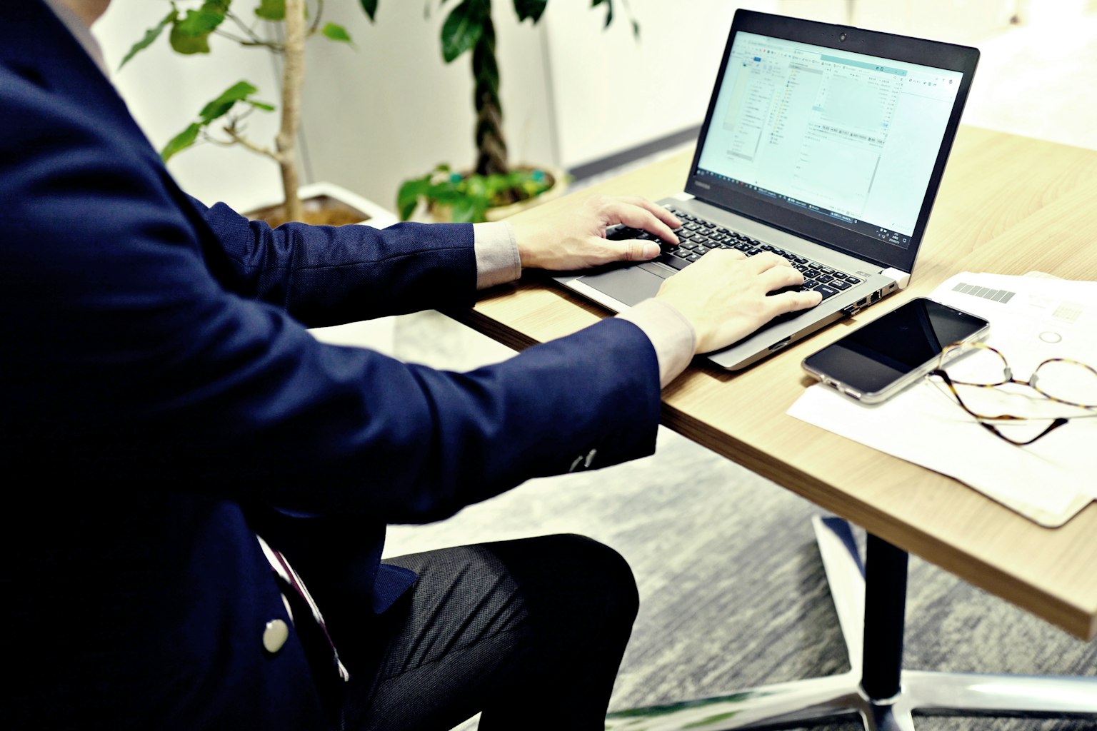 Ein Geschäftsmann, der an einem Schreibtisch auf einem Laptop tippt, mit einem Smartphone und einer Brille in der Nähe