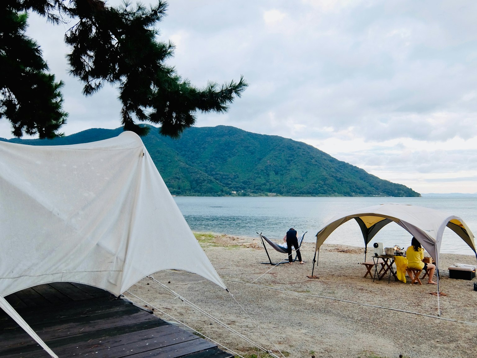 Pemandangan pantai dengan tenda, gunung, dan langit mendung di latar belakang