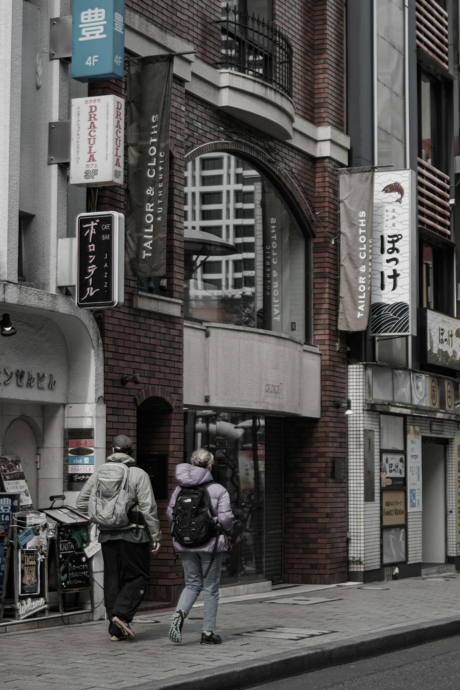 Two people walking on the street beside a building with various signs