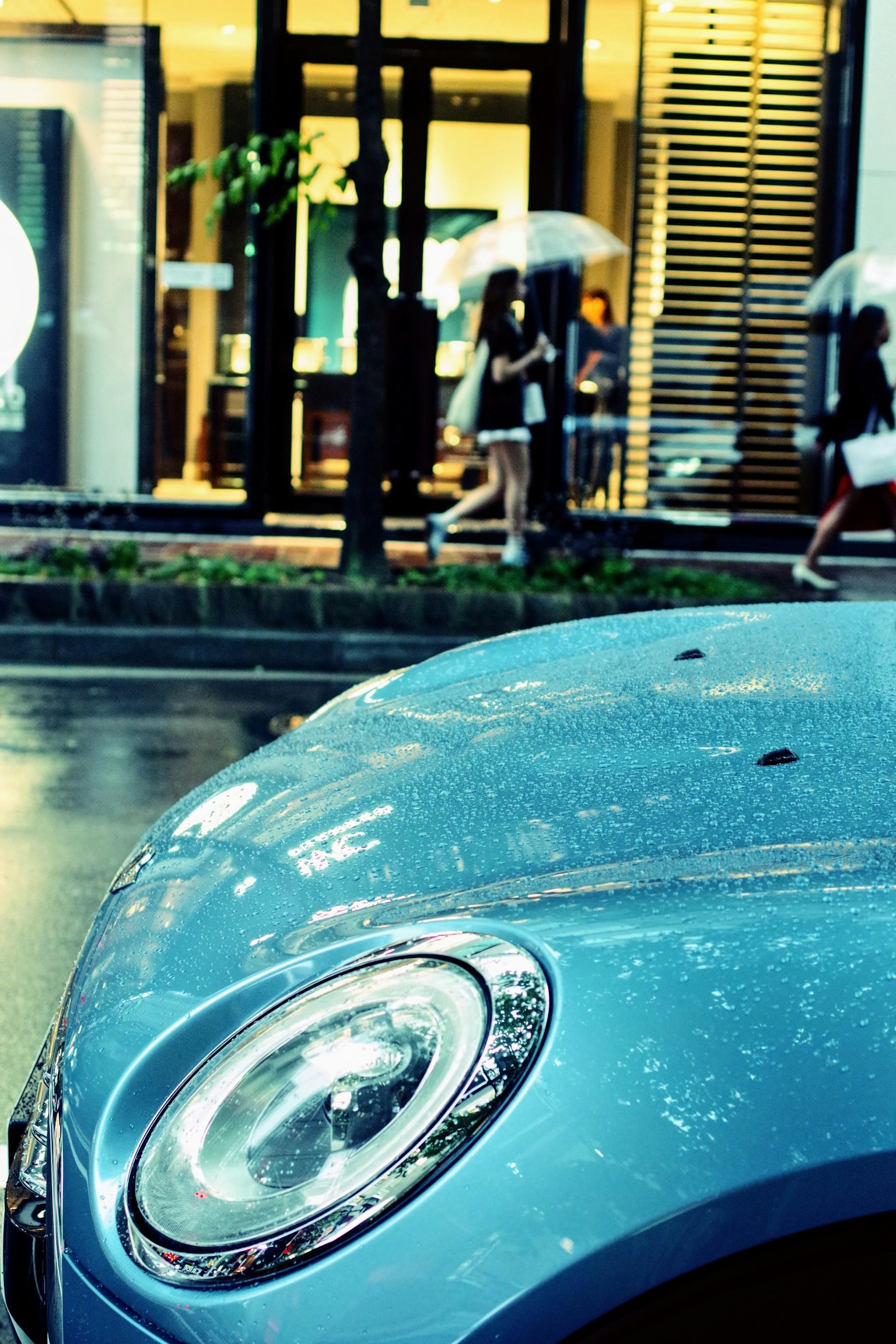 Blue car with raindrops and pedestrians holding umbrellas