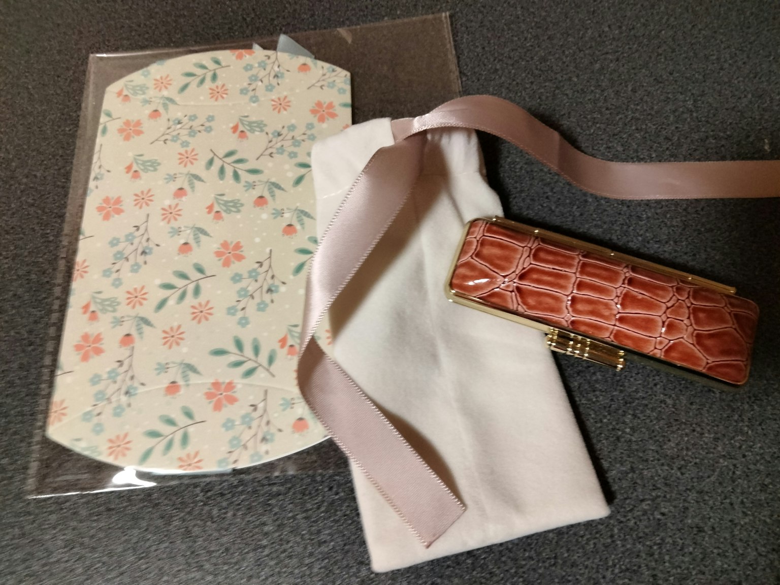 Une enveloppe à motif floral avec des fleurs roses et des feuilles vertes à côté d'un sac en tissu blanc et d'un étui en crocodile doré