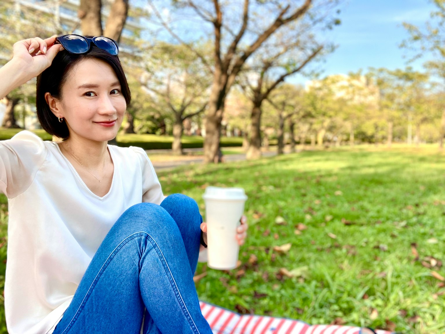 공원에서 커피를 들고 있는 여성, 파란 청바지와 흰색 티셔츠를 입고, 손에 선글라스를 들고 있음