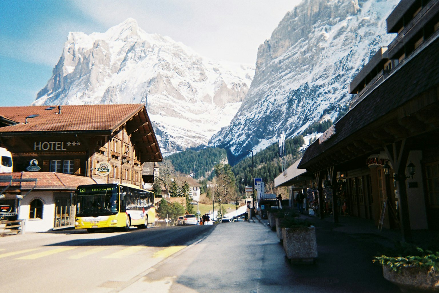 Scena di villaggio con montagne innevate sullo sfondo un autobus che percorre la strada e edifici in legno tradizionali lungo la strada