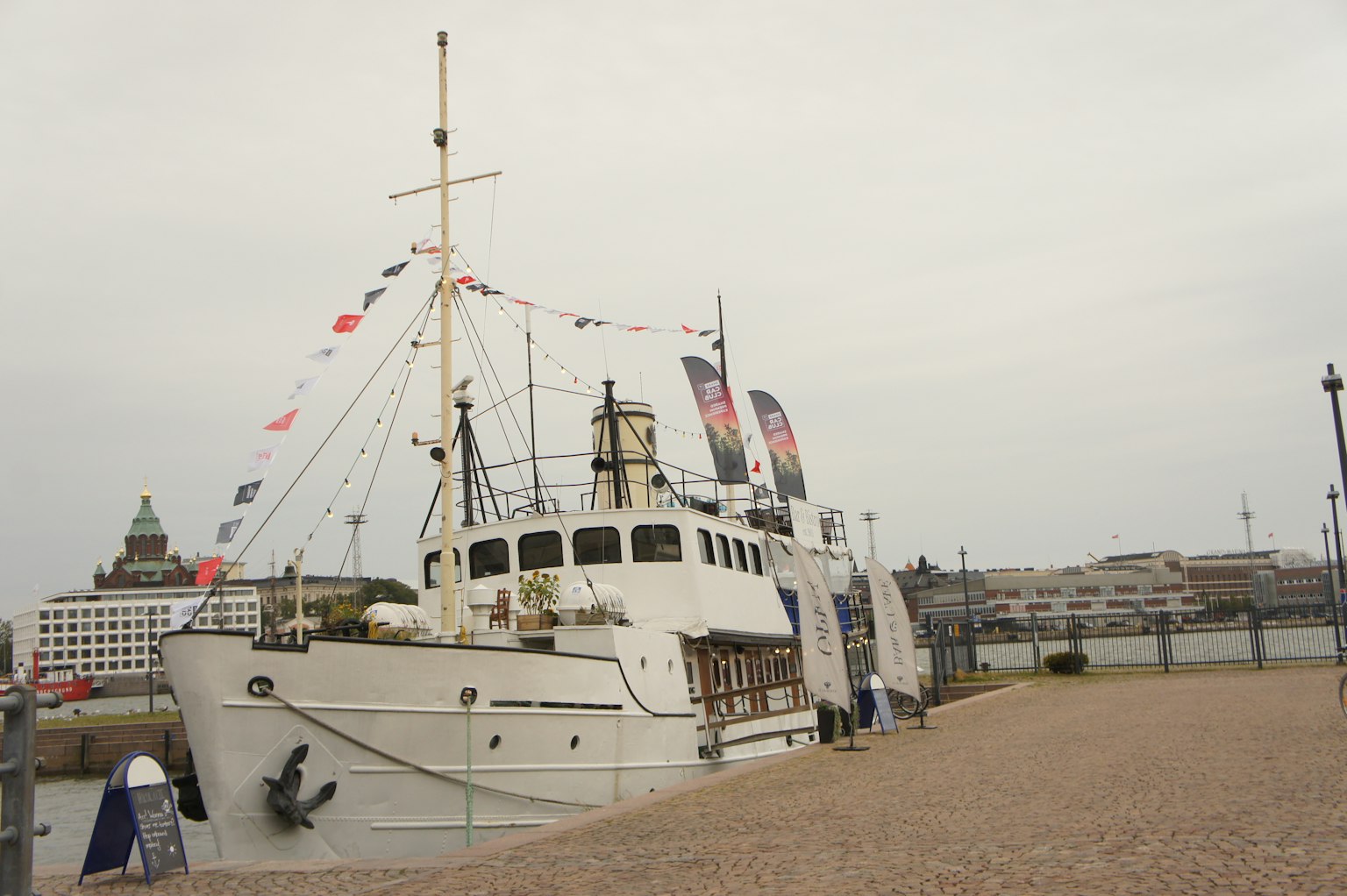 Un barco blanco atracado en la orilla con banderas decorativas y un paisaje urbano al fondo