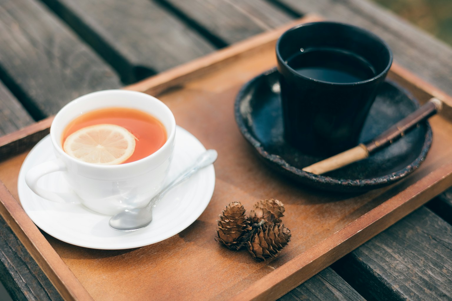 Una taza blanca con una rodaja de limón flotando en té y una taza negra de café colocadas sobre una bandeja de madera