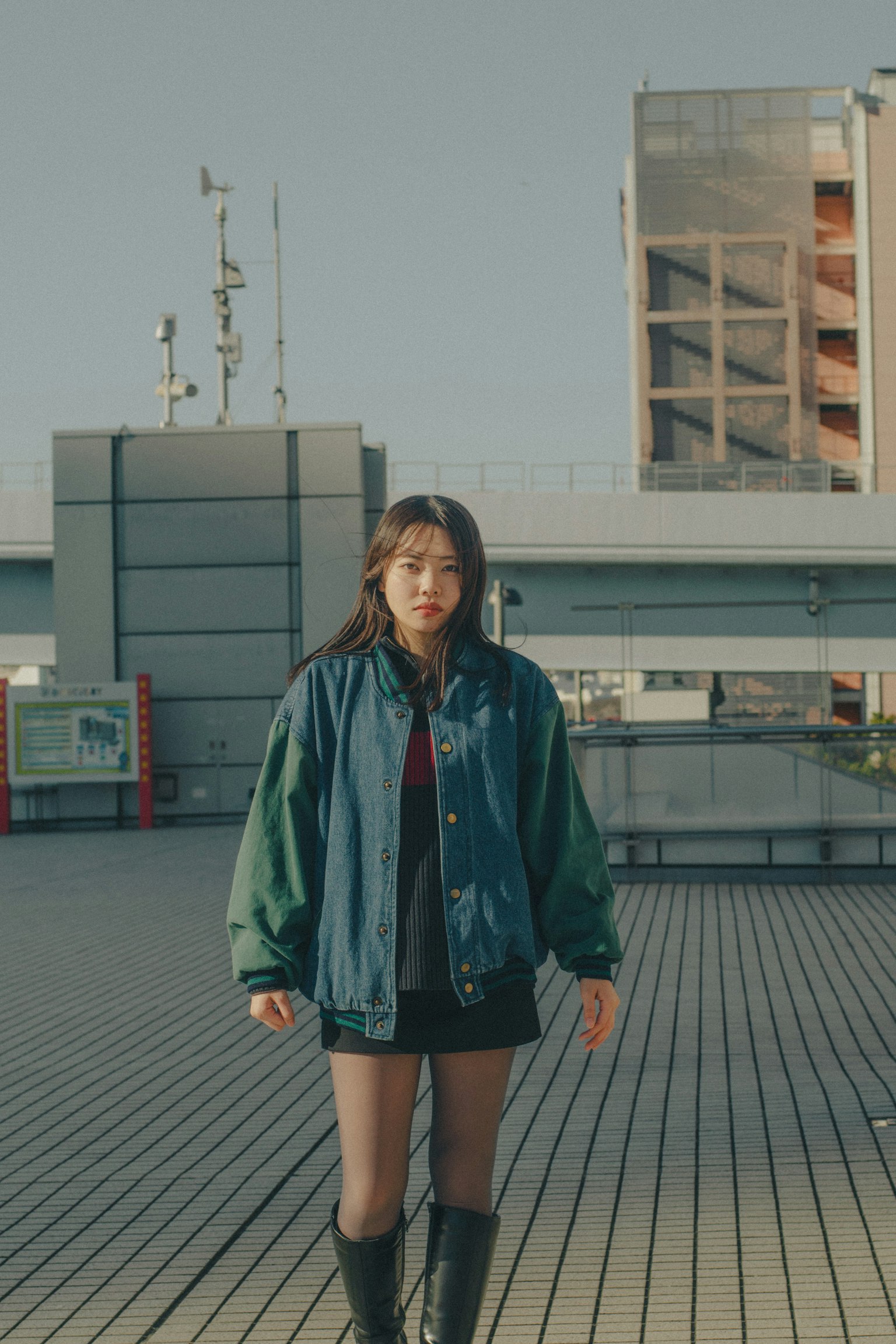 一位女性在戶外站立，穿著休閒街頭風格的服裝，搭配綠色和藍色的夾克以及黑色短裙