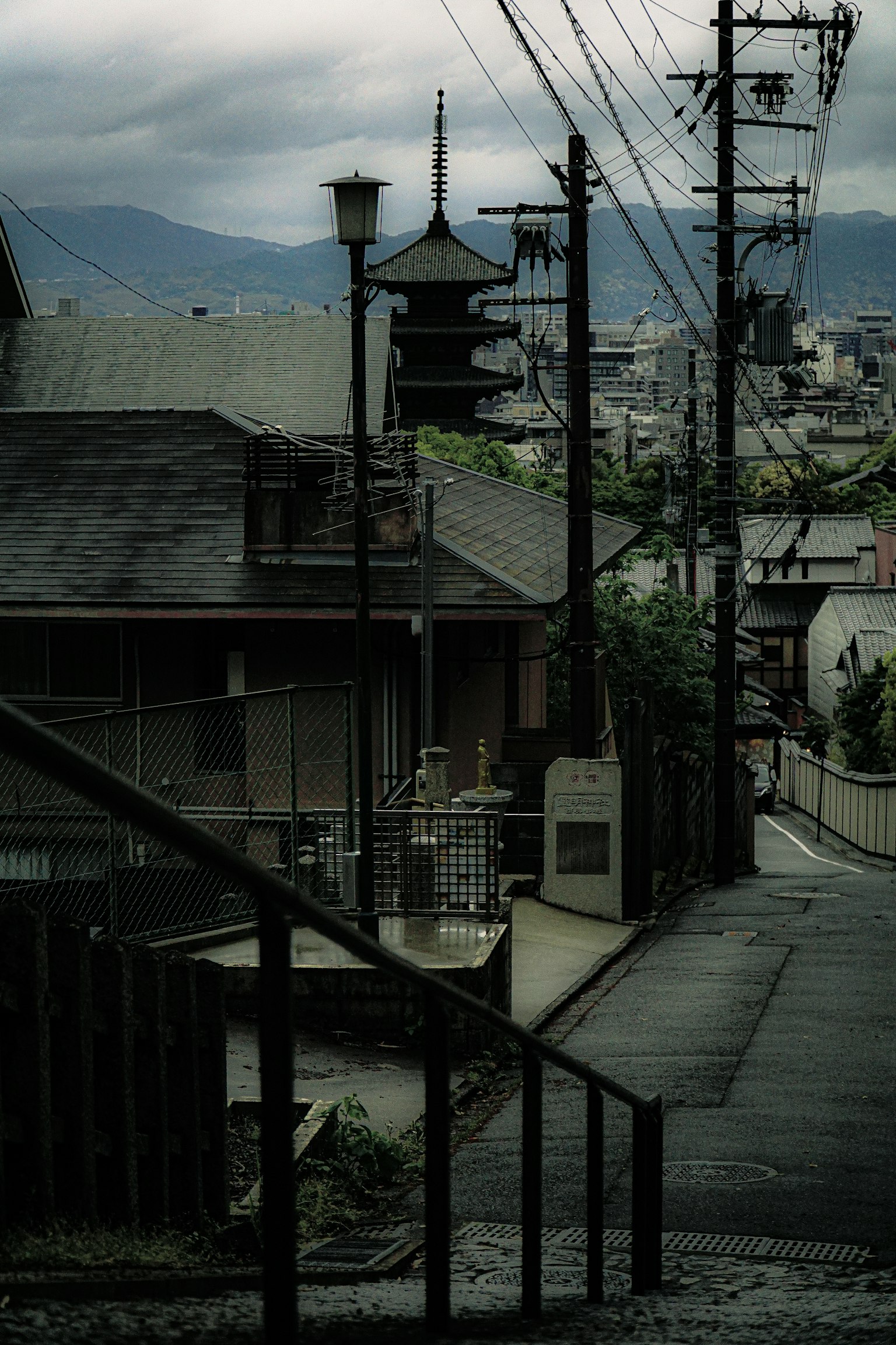 Japanese residential street under a cloudy sky featuring a pagoda-like structure in the distance and utility poles