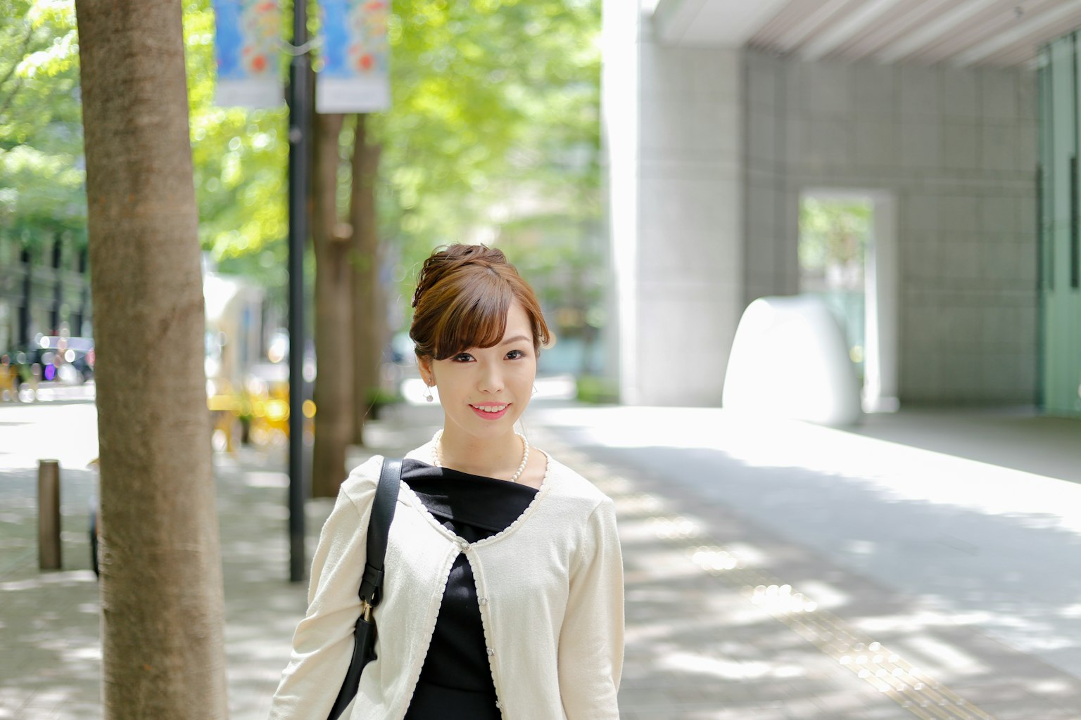 Portrait of a smiling woman in a park wearing a white cardigan with green trees and a modern building in the background