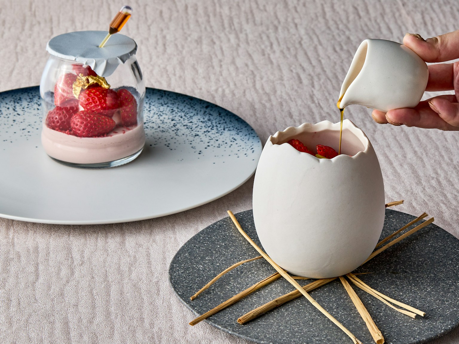 A hand pouring sauce into a white egg-shaped dessert with a fruit dessert in a jar beside it