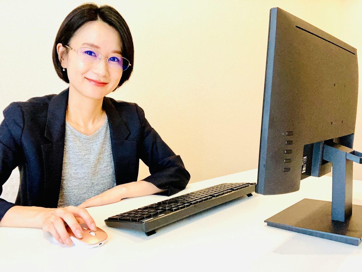一位女性坐在電腦前，穿著灰色T恤和西裝外套，佩戴眼鏡