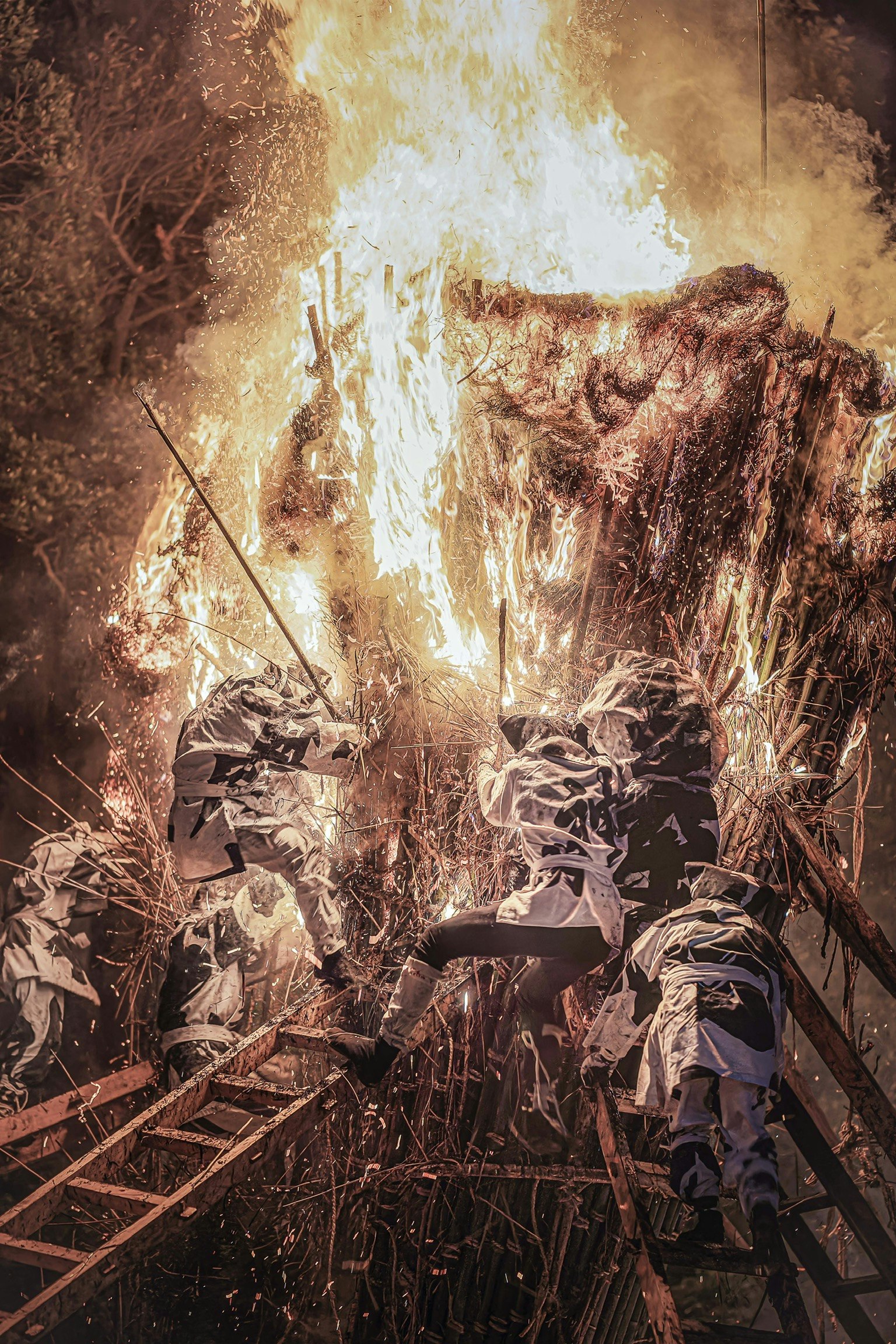 炎に包まれた祭りのシーンで、白い衣装を着た人々が火の近くで活動している