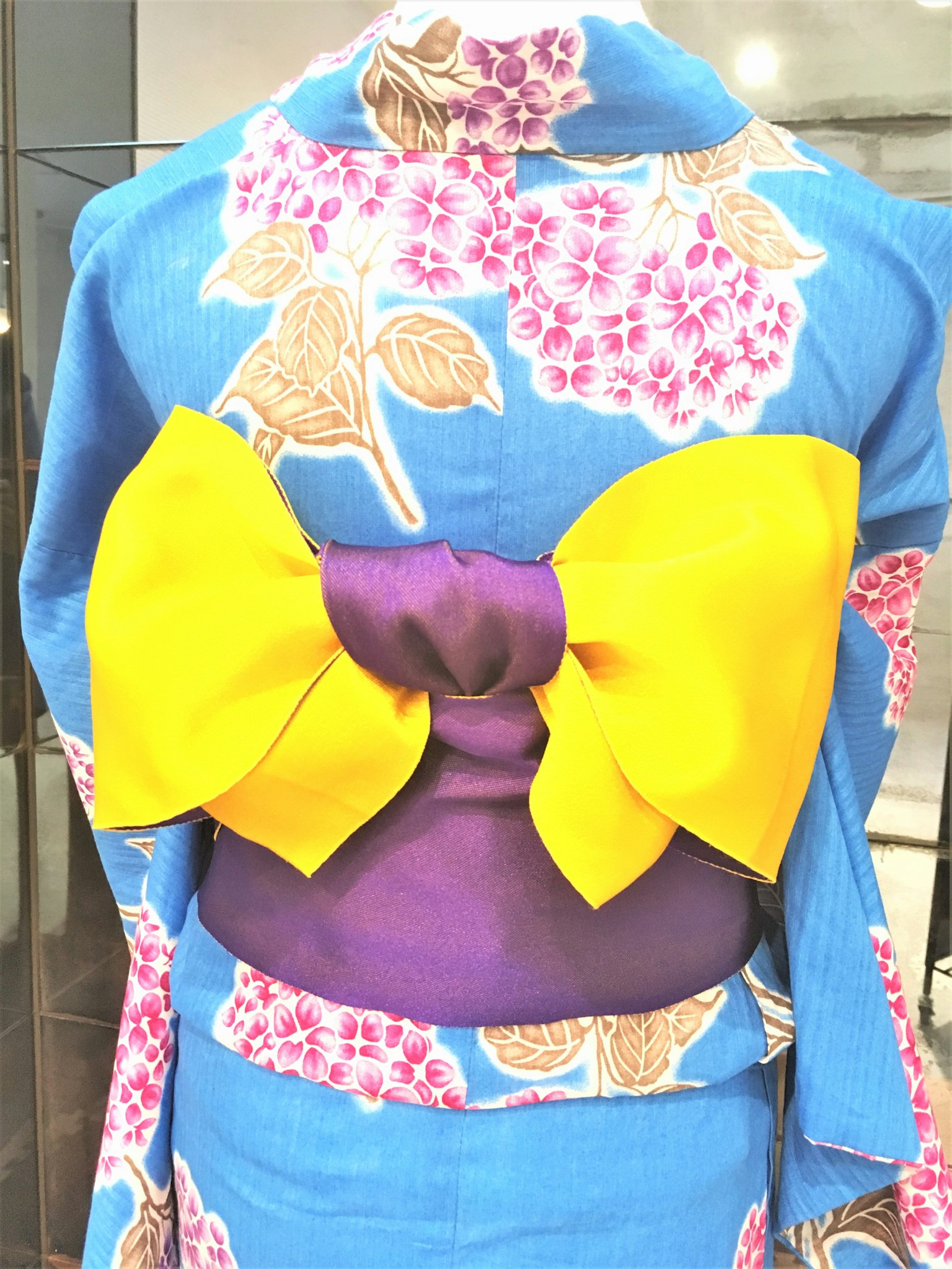 Kimono biru dengan pola bunga menampilkan pita kuning besar dan obi ungu