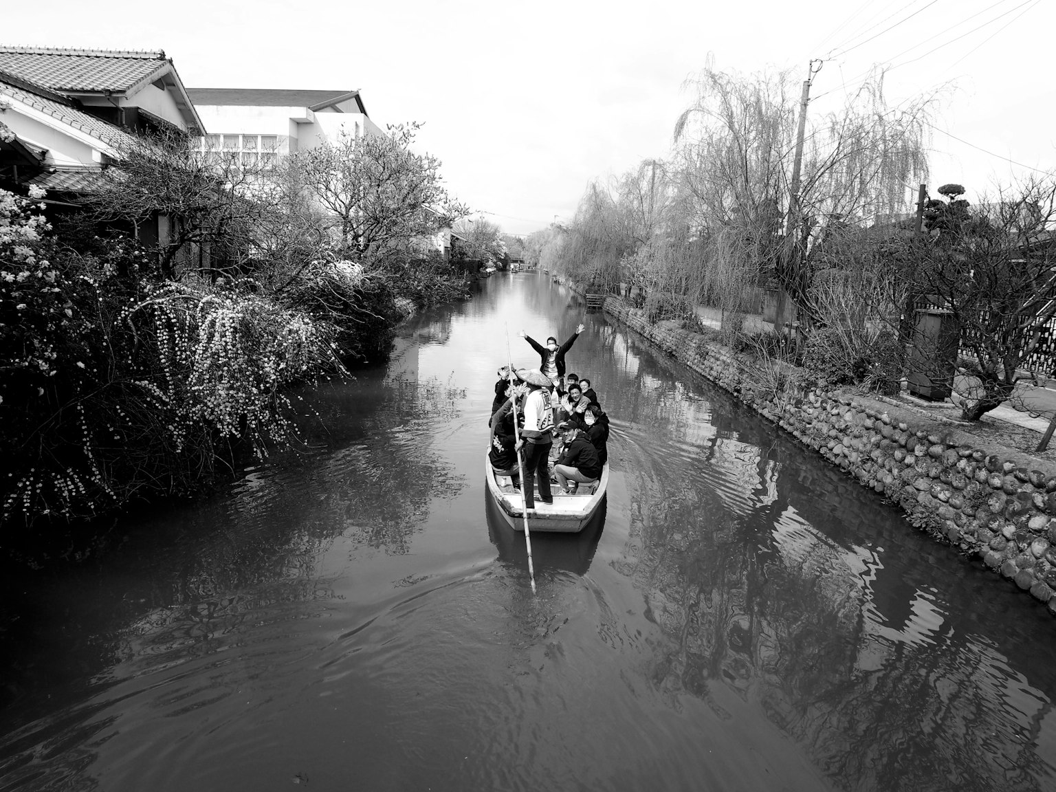 ภาพขาวดำของเรือเล็กที่แล่นอยู่ในคลองที่ถูกล้อมรอบด้วยต้นไม้และบ้านเรือน