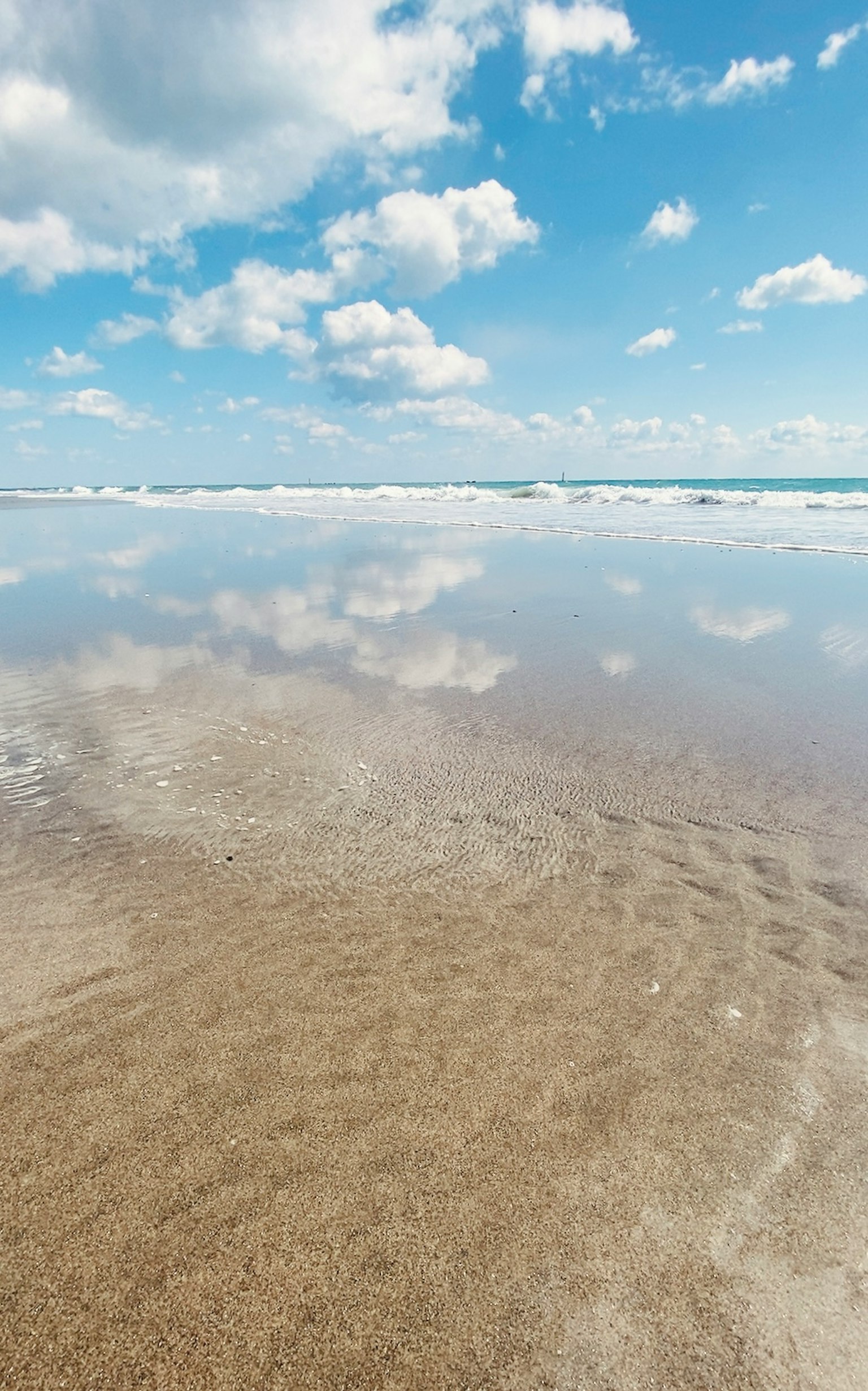 Pemandangan pantai tenang dengan refleksi awan putih di langit biru di atas air