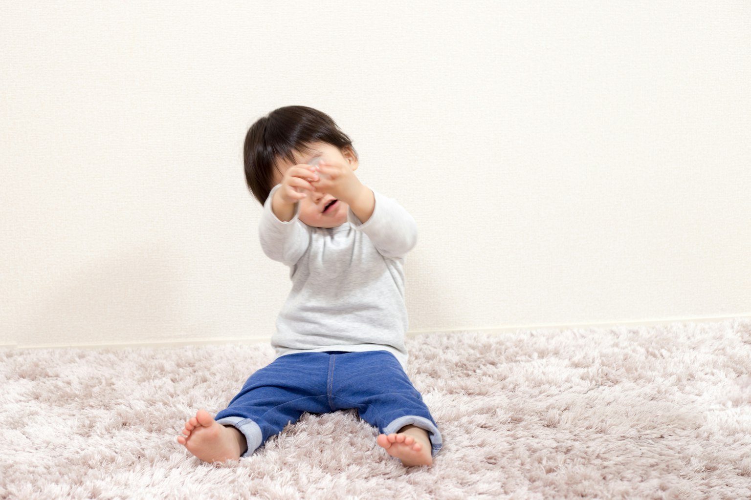 Un bambino seduto su un tappeto morbido davanti a un muro bianco che si copre il viso con le mani