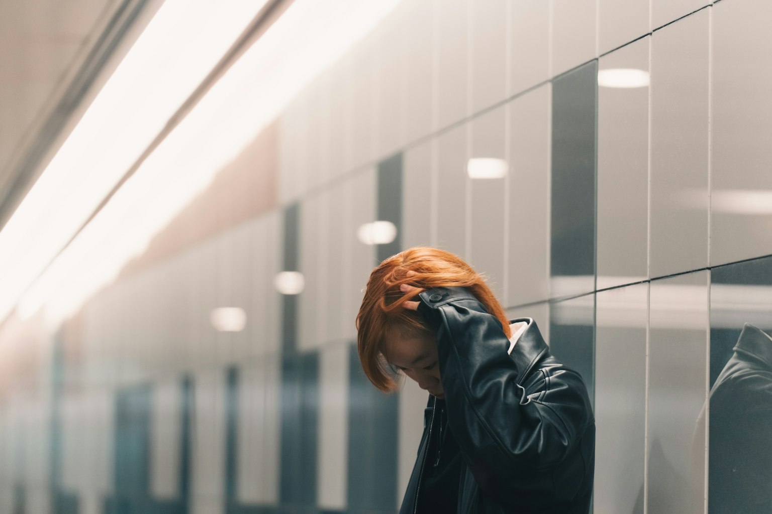 Personne avec des cheveux orange touchant ses cheveux en se tenant devant un mur portant une veste noire éclairage argenté réfléchissant