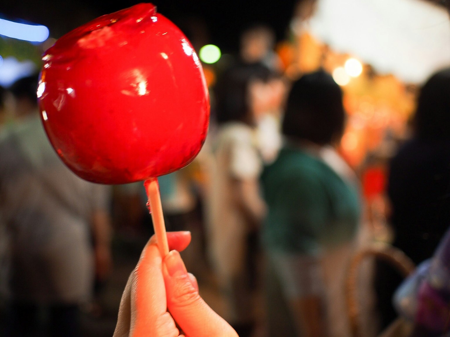A hand holding a bright red candy apple with a blurred crowd in the background