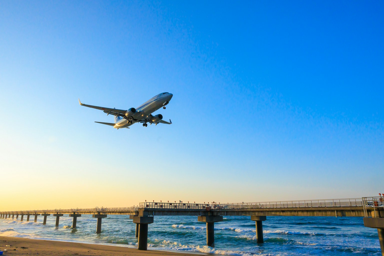 Flugzeug fliegt über einen Pier mit Ozeanwellen unter blauem Himmel