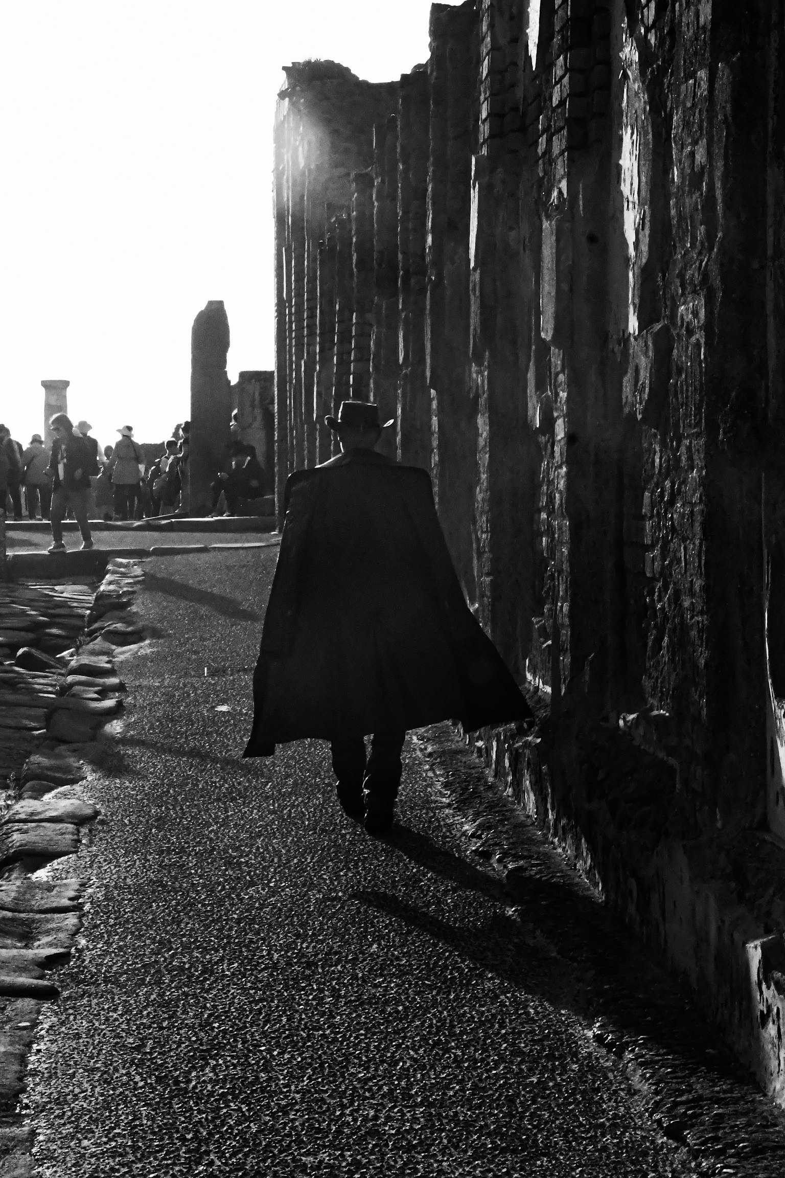 黒いコートを着た人物が古い壁の前を歩いているモノクロの風景