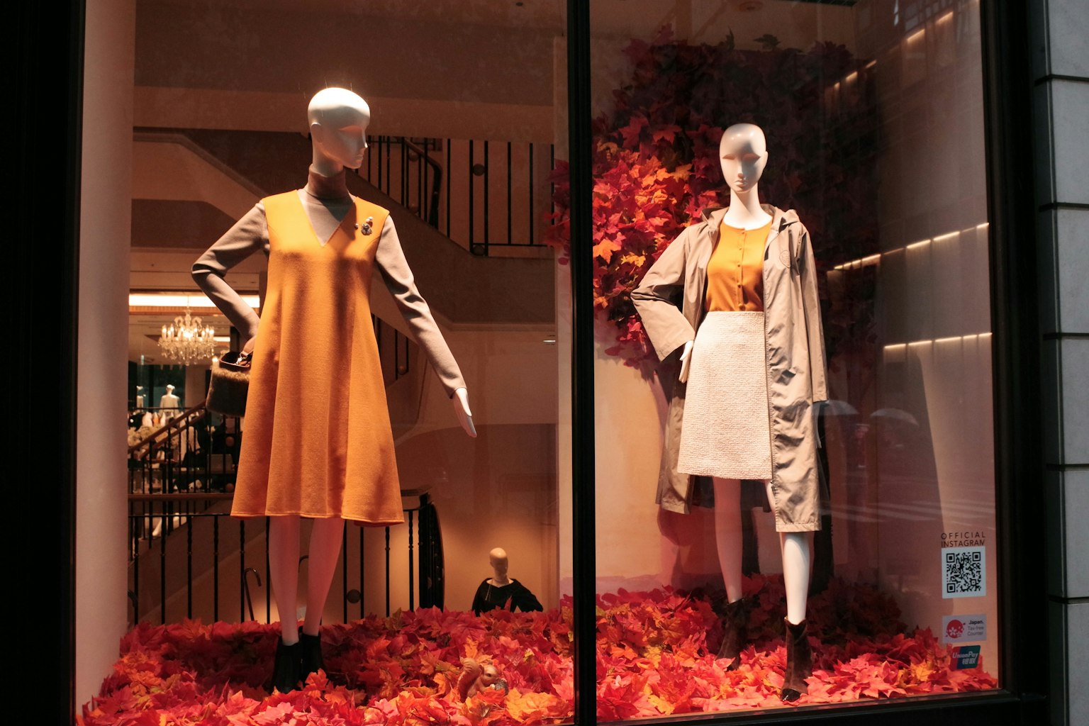 Deux mannequins dans une vitrine portant une robe orange et une robe blanche entourés de feuilles d'automne